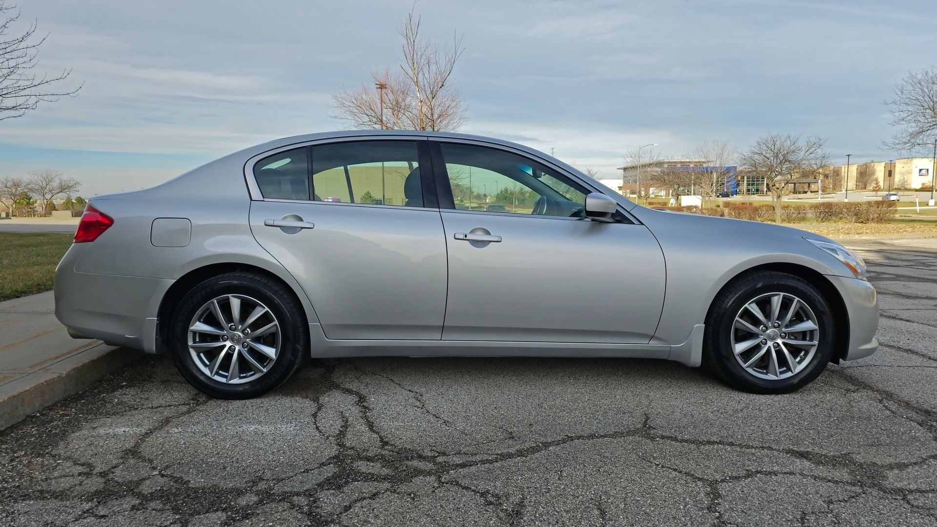 Silver sedan parked on asphalt next to a curb. Sunny day, with trees and a building in the background.