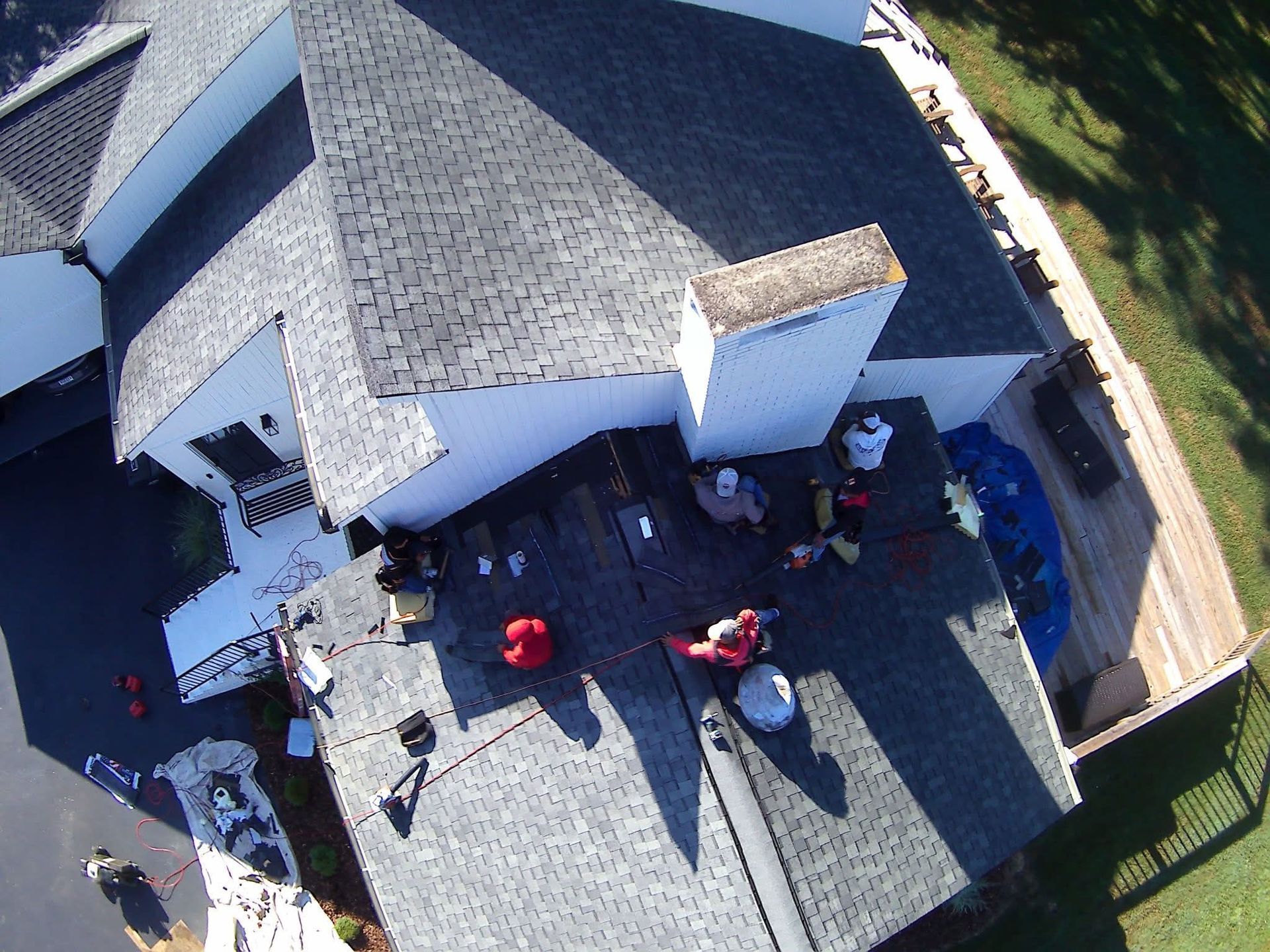An aerial view of roofers in red shirts working on the shingled roof of a house next to a white chimney and a wooden deck.