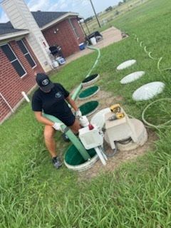 Person kneeling by outdoor utility tanks, handling hoses beside a house in a grassy yard.