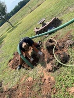 Dog standing by a green hose on muddy ground in a grassy yard, with a log stump nearby.