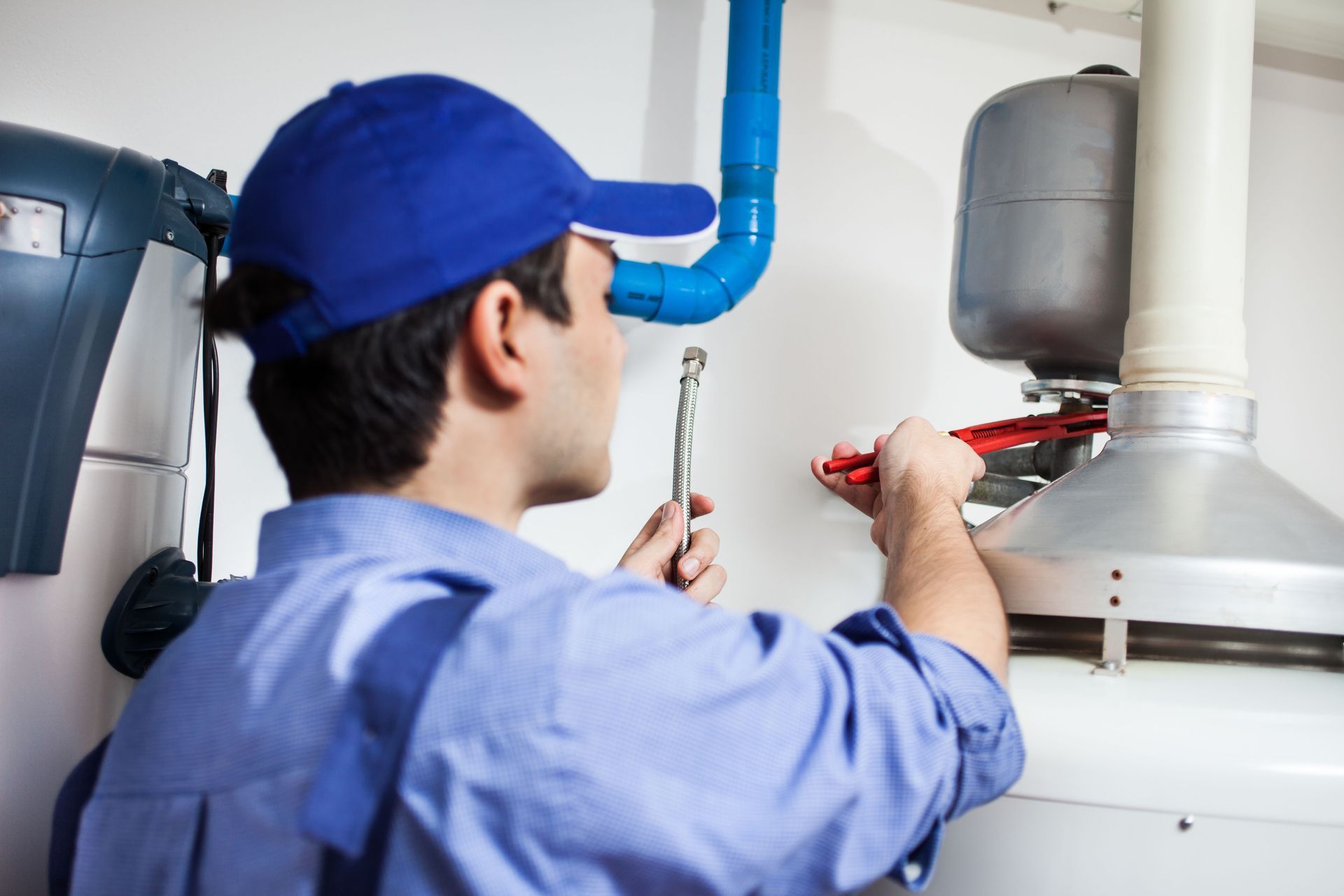 Plumber in blue shirt and cap, using a wrench to work on a water heater.