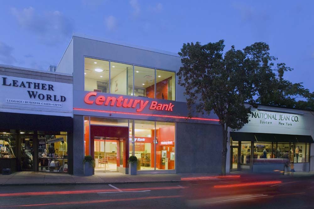 Century Bank building with red neon sign, flanked by shops, at dusk.