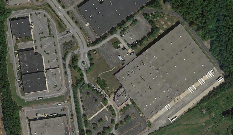 Aerial view of industrial buildings, parking lots, and roads; a large warehouse dominates the right side.