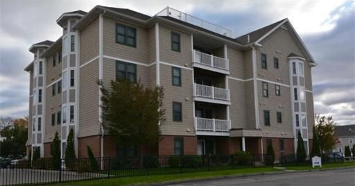 Multi-story beige apartment building with white balconies and brick base under a cloudy sky.