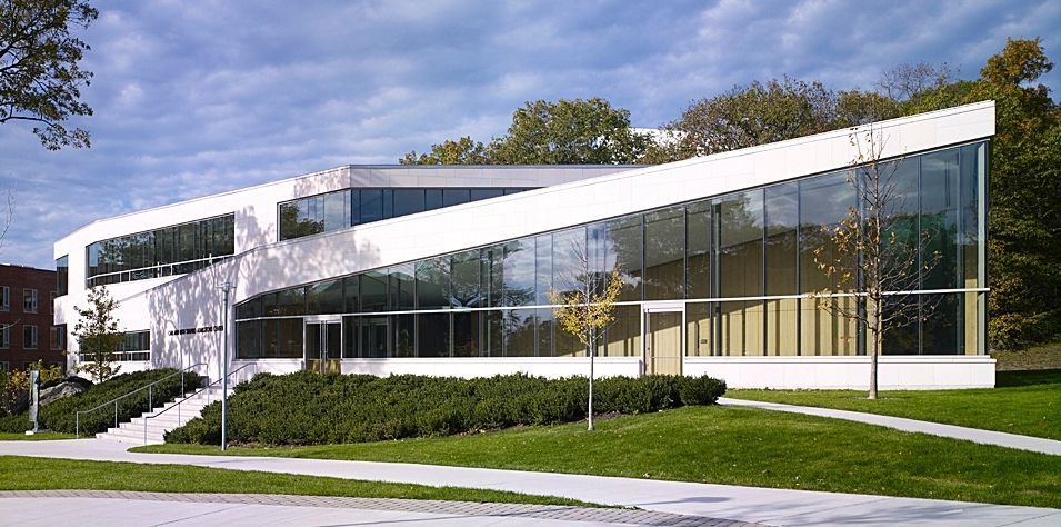 Modern white building with angled roof and glass walls, set on a grassy lawn with trees in the background.