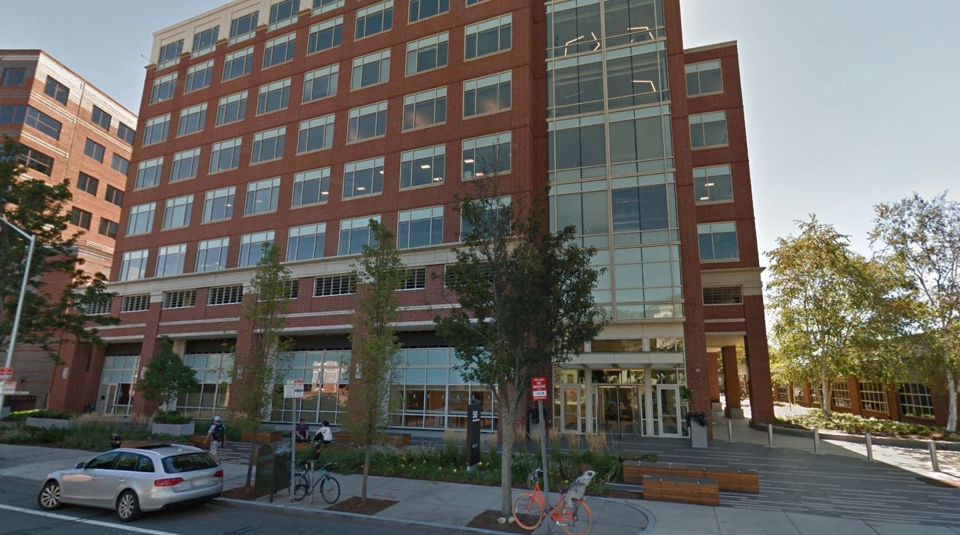 Brick building with glass entrance; people near the doors. Silver car parked on the street in front.