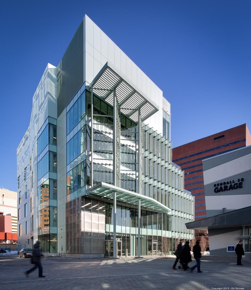 Modern glass-walled building with pedestrian entrance, passersby on a paved street. Sunny day.