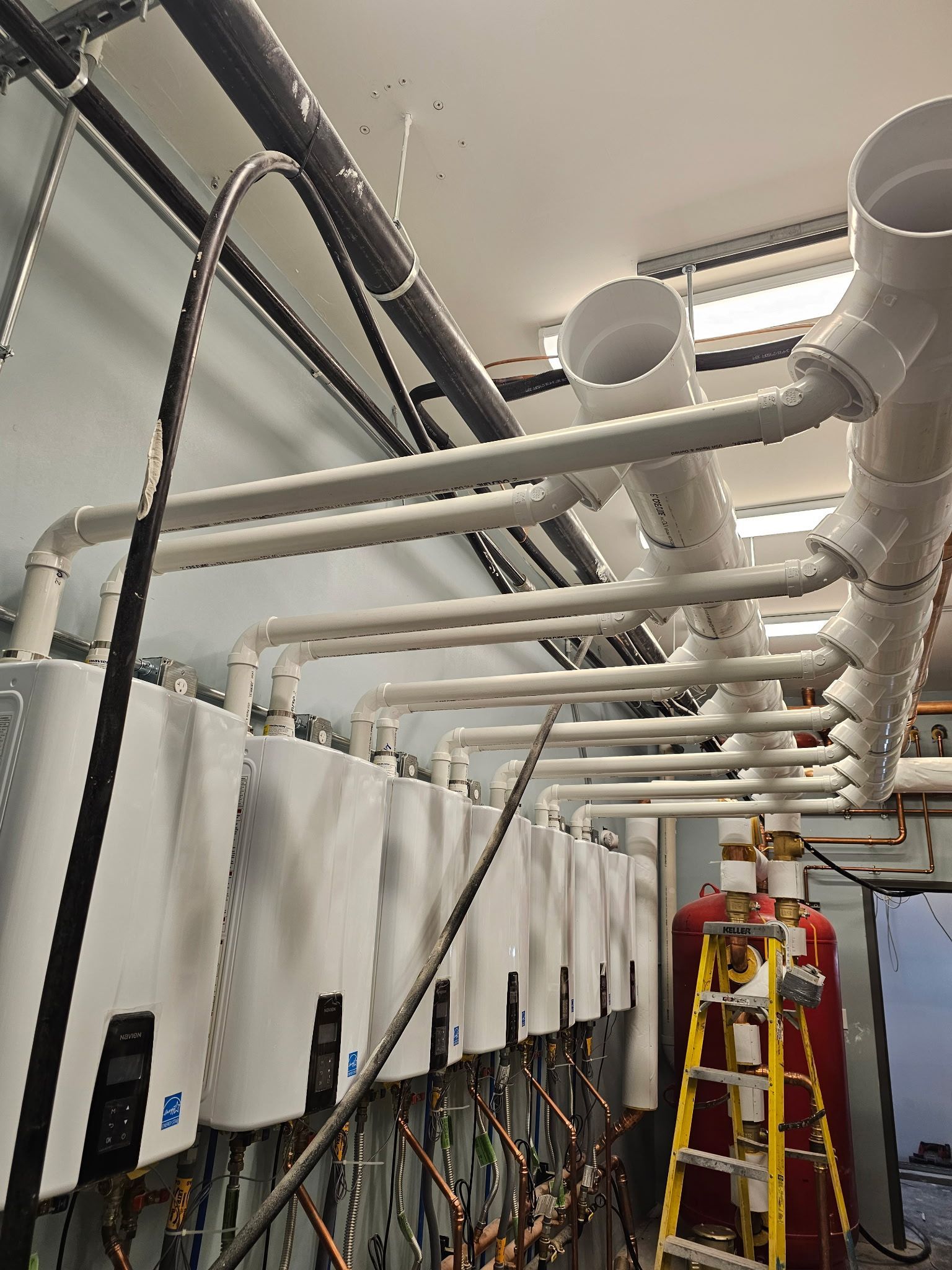 Row of white water heaters with connected white pipes in a utility room.
