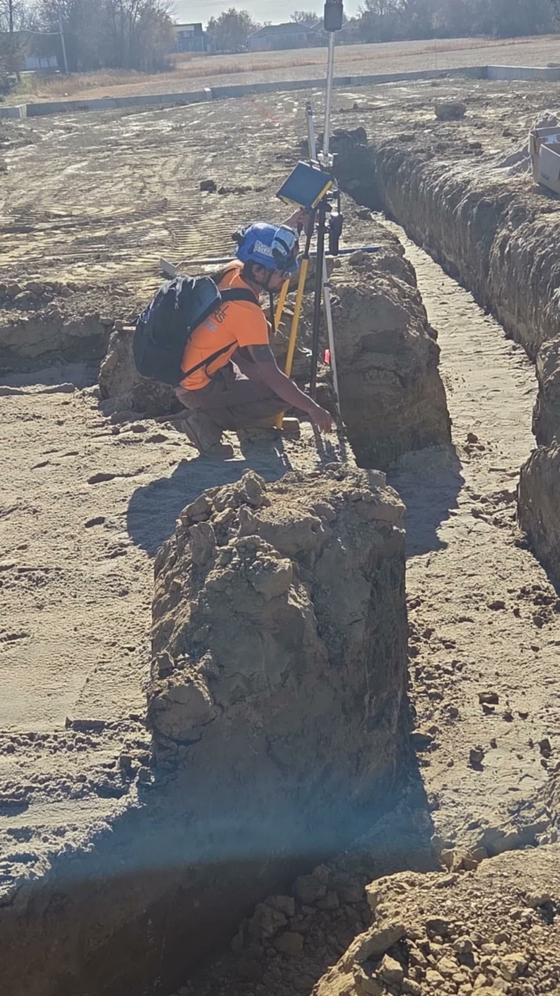 Worker in orange vest kneels in a trench, surveying with equipment; dirt field, sunny day.
