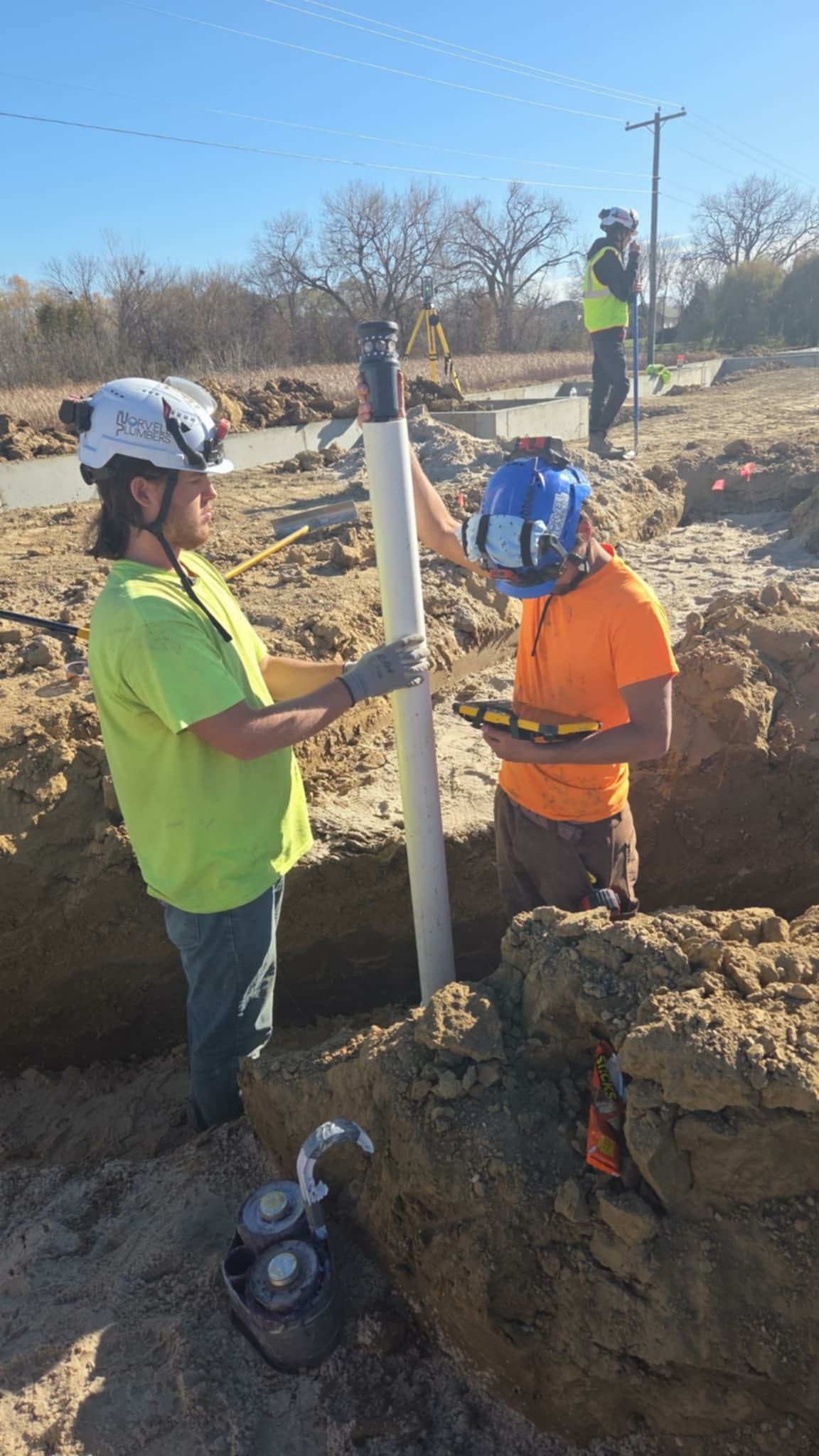 Two workers in hard hats install a pipe in a trench; a third worker watches in the background on a sunny day.
