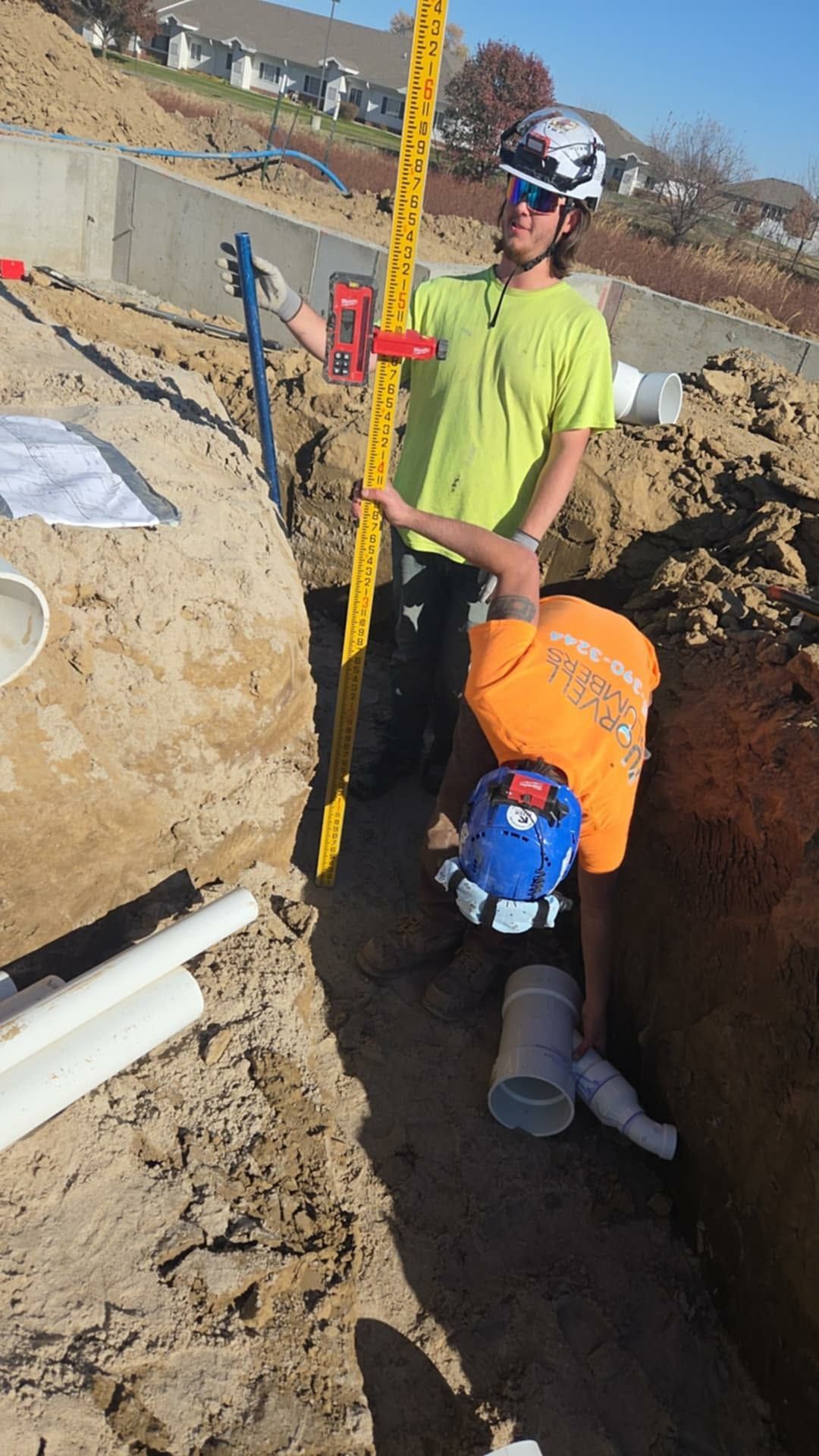 Two construction workers checking pipe in a trench with level and measuring stick; sunny day.