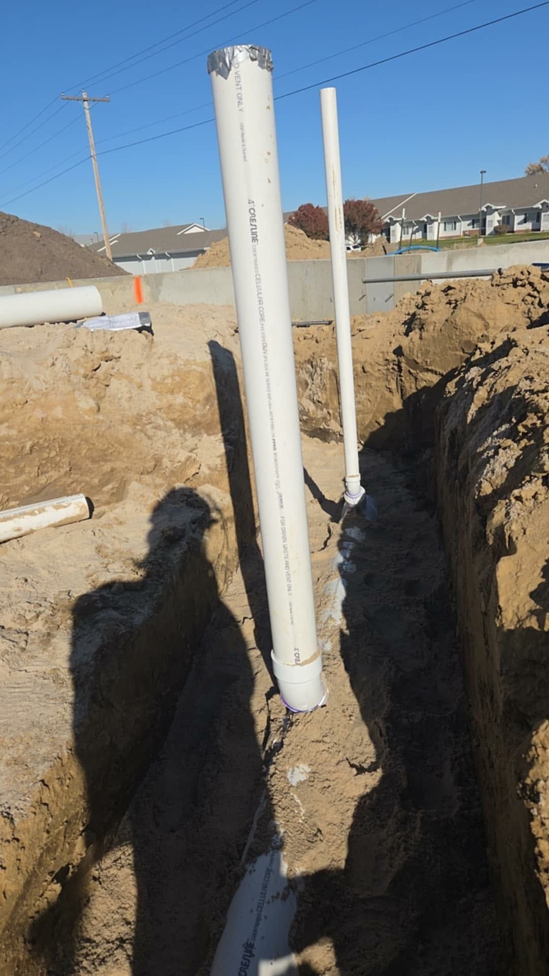 Construction trench with PVC pipes in the ground, sunlight, blue sky, and a suburban backdrop.