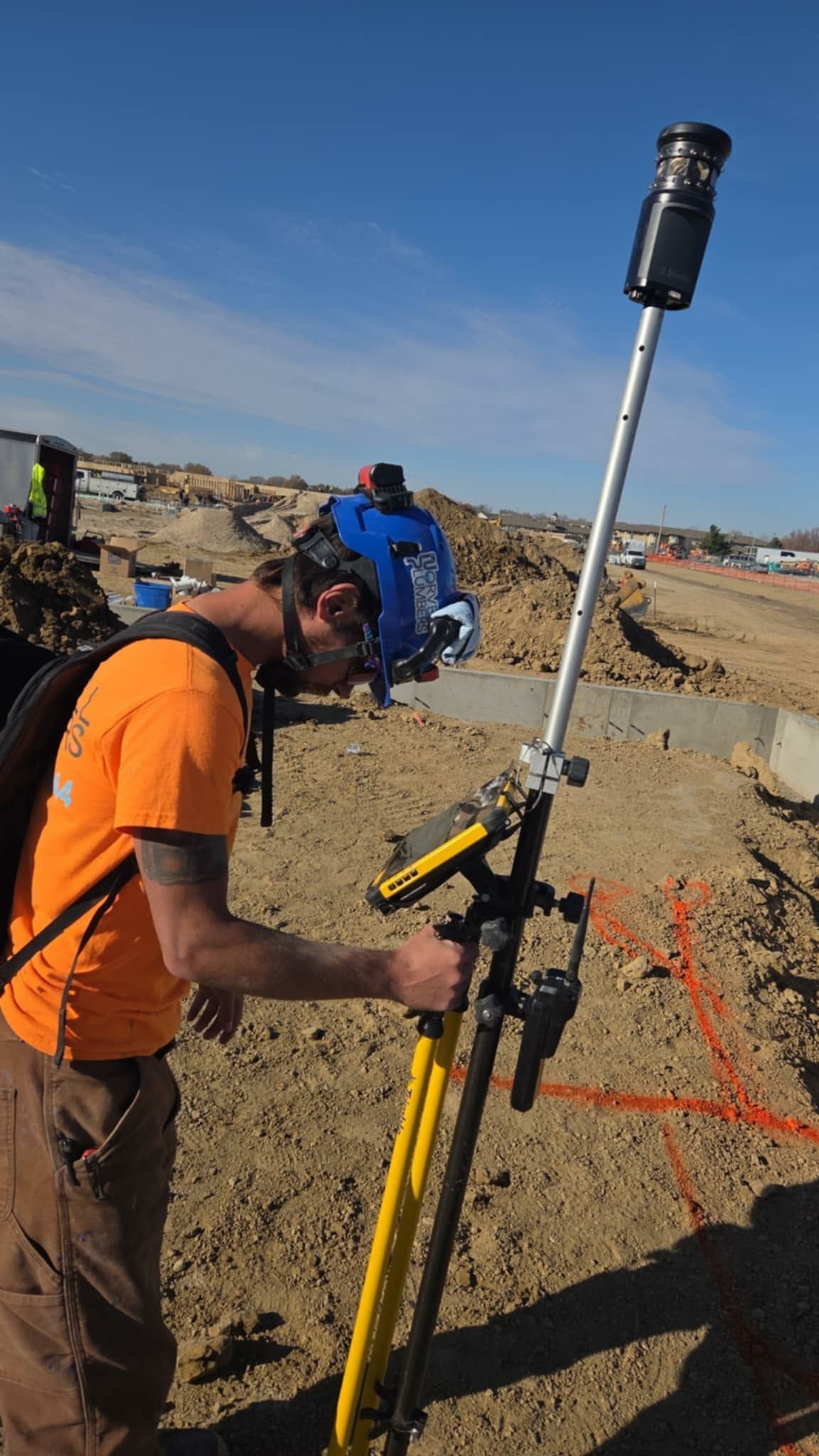 Construction worker using surveying equipment on a construction site; clear blue sky in background.