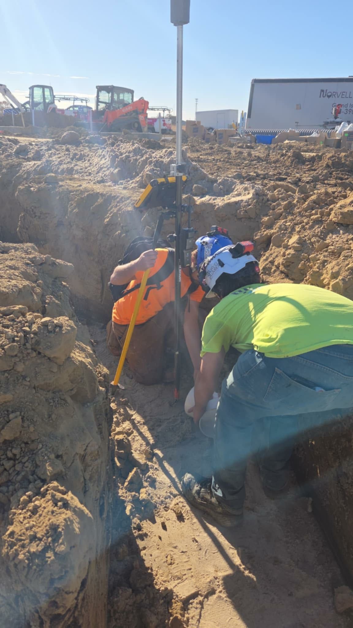 Two workers in a trench; one uses a tool, one holds a device. Bright sun, construction site.