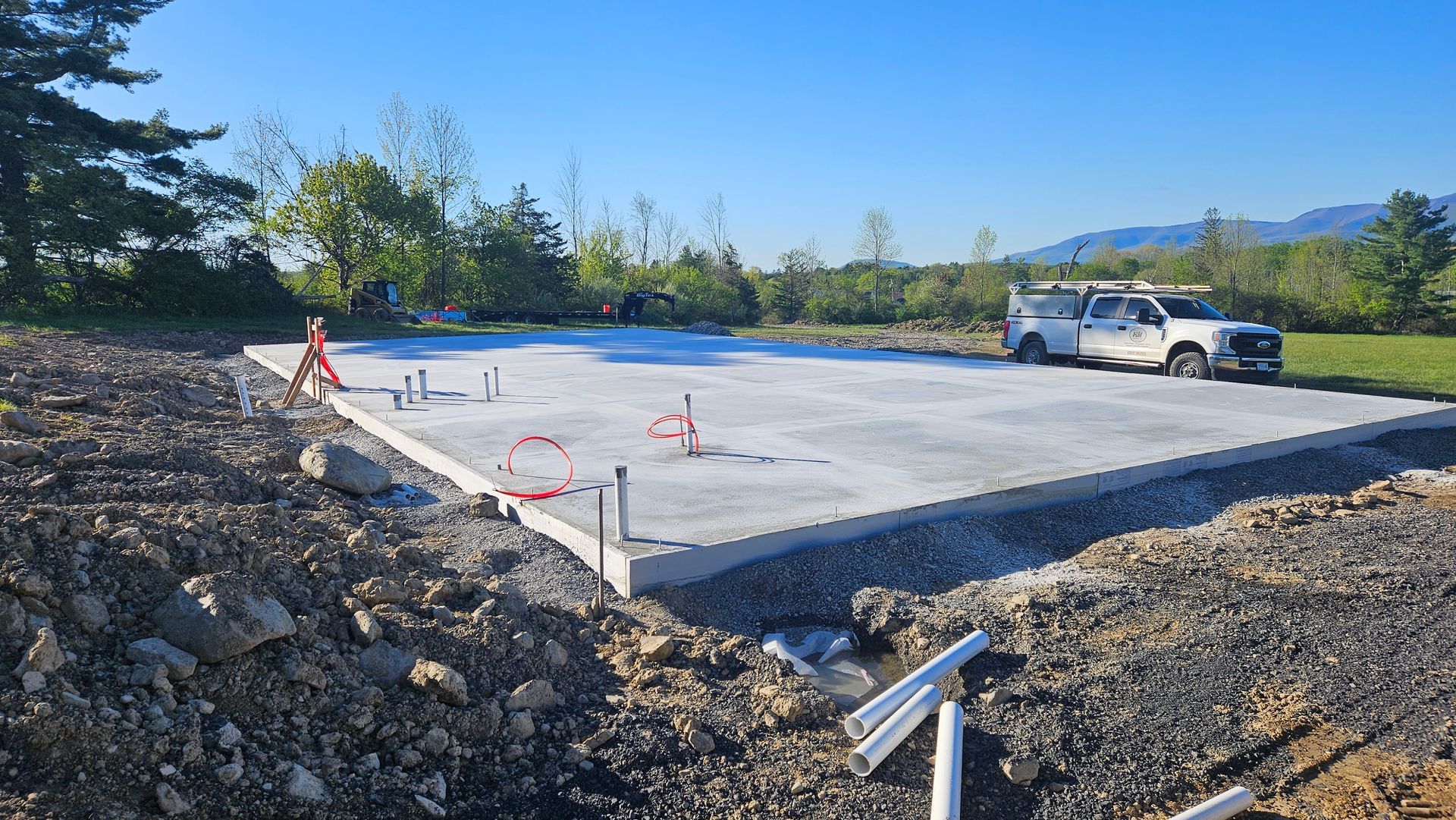 Concrete foundation slab at a construction site with pipes and a white truck nearby, under a blue sky