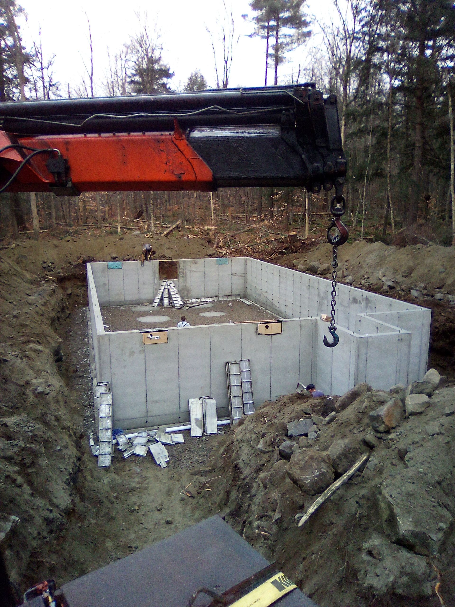Foundation walls being built at a wooded construction site, with an excavator arm overhead.