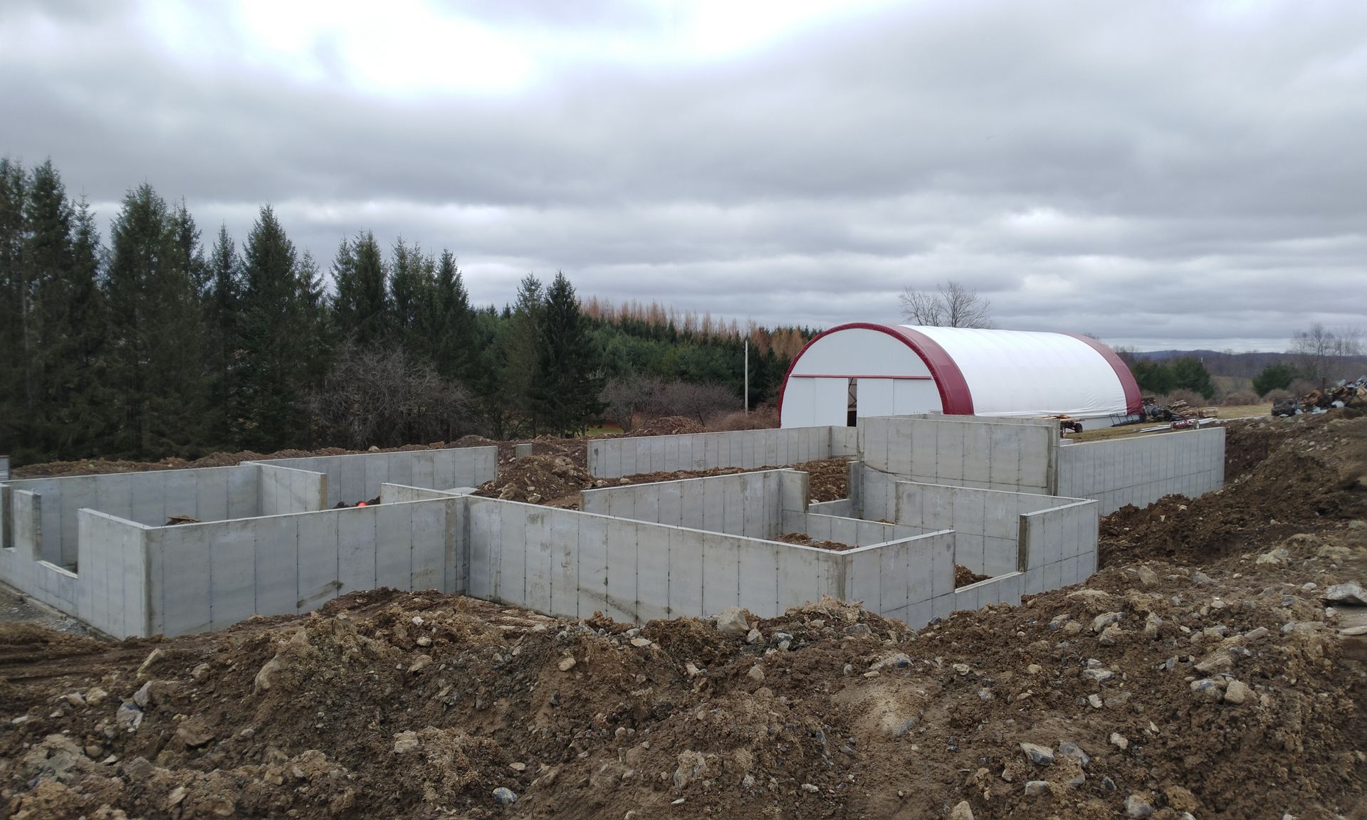 Concrete foundation walls under construction beside a white-and-red quonset hut on a cloudy rural site