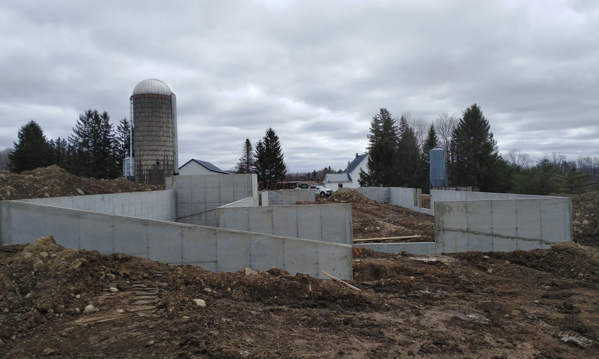 Partially built farm structure with concrete walls, dirt foreground, silo, and cloudy sky