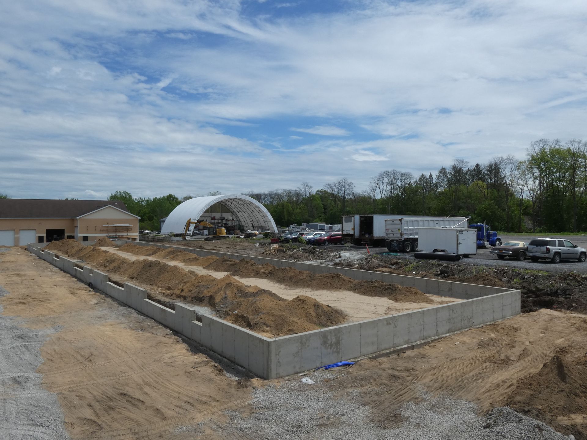 Construction site with concrete foundation walls, dirt piles, trucks, and a white arched tent under a cloudy sky