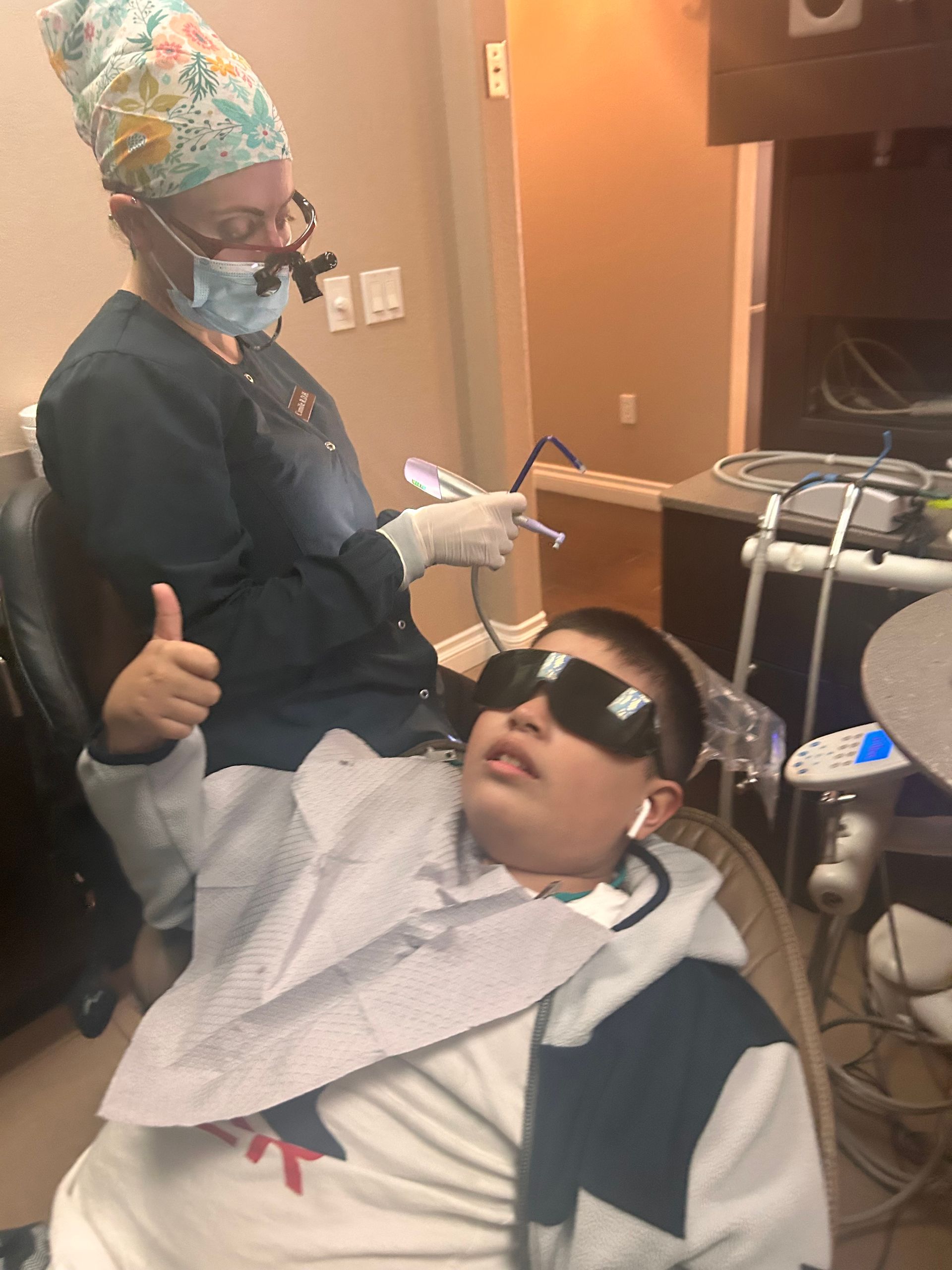 A dental professional prepares equipment while a patient in dark protective glasses gives a thumbs-up in a dental chair.