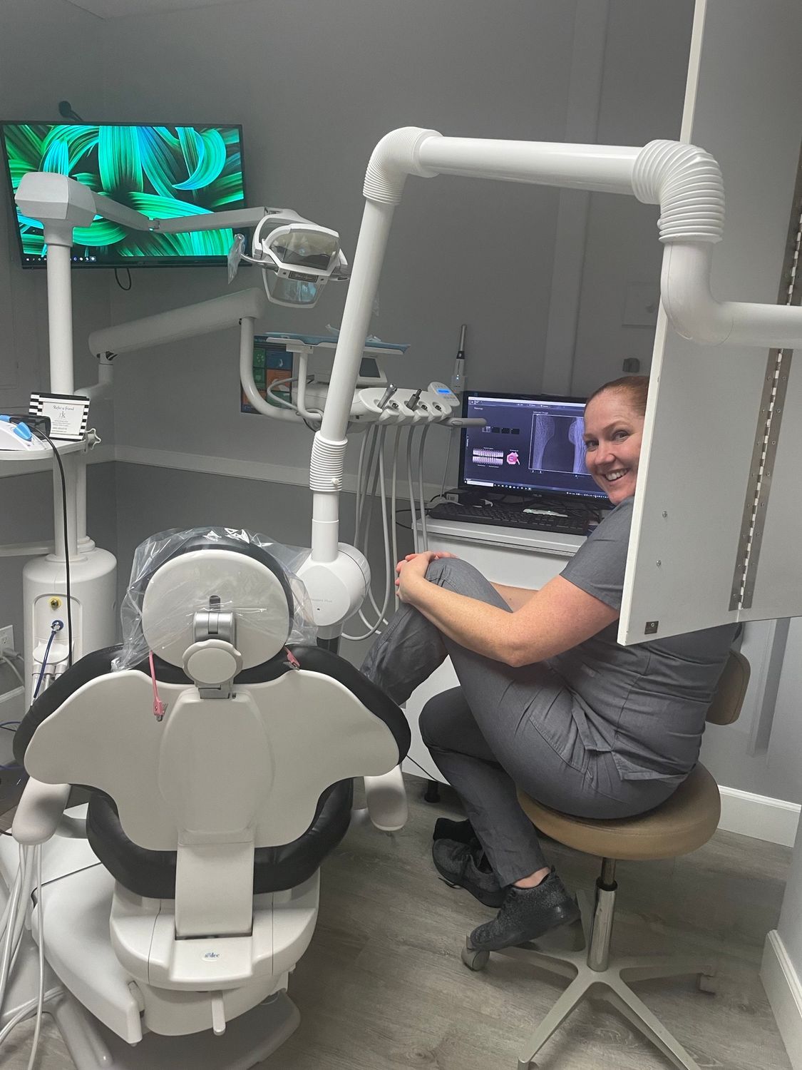 A professional in scrubs sits on a rolling stool in a dental exam room, smiling toward the camera near a computer screen.