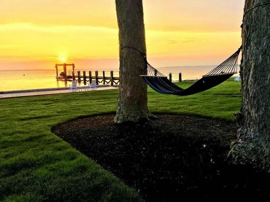 A hammock hangs between two trees on a grassy lawn overlooking a lake at sunset with a pier in the distance.