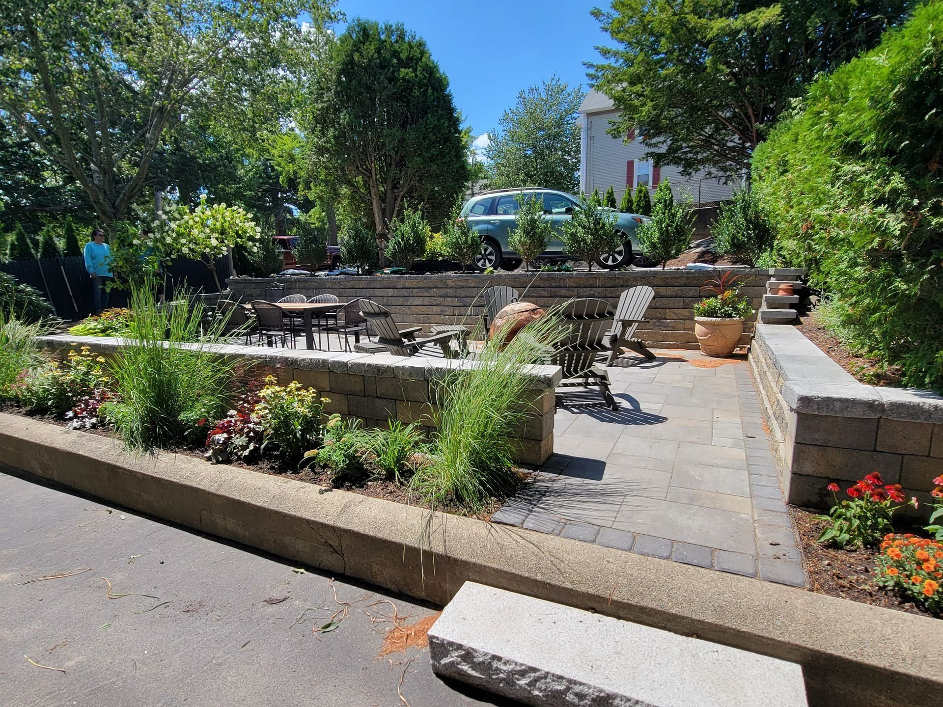 A tiered stone patio with outdoor furniture, landscaping, and a car parked on an upper level, seen from a paved driveway.