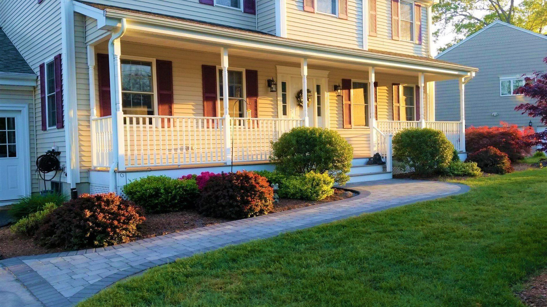 A suburban house with a tan exterior, dark shutters, and a front porch with a white railing and walkway at sunset.