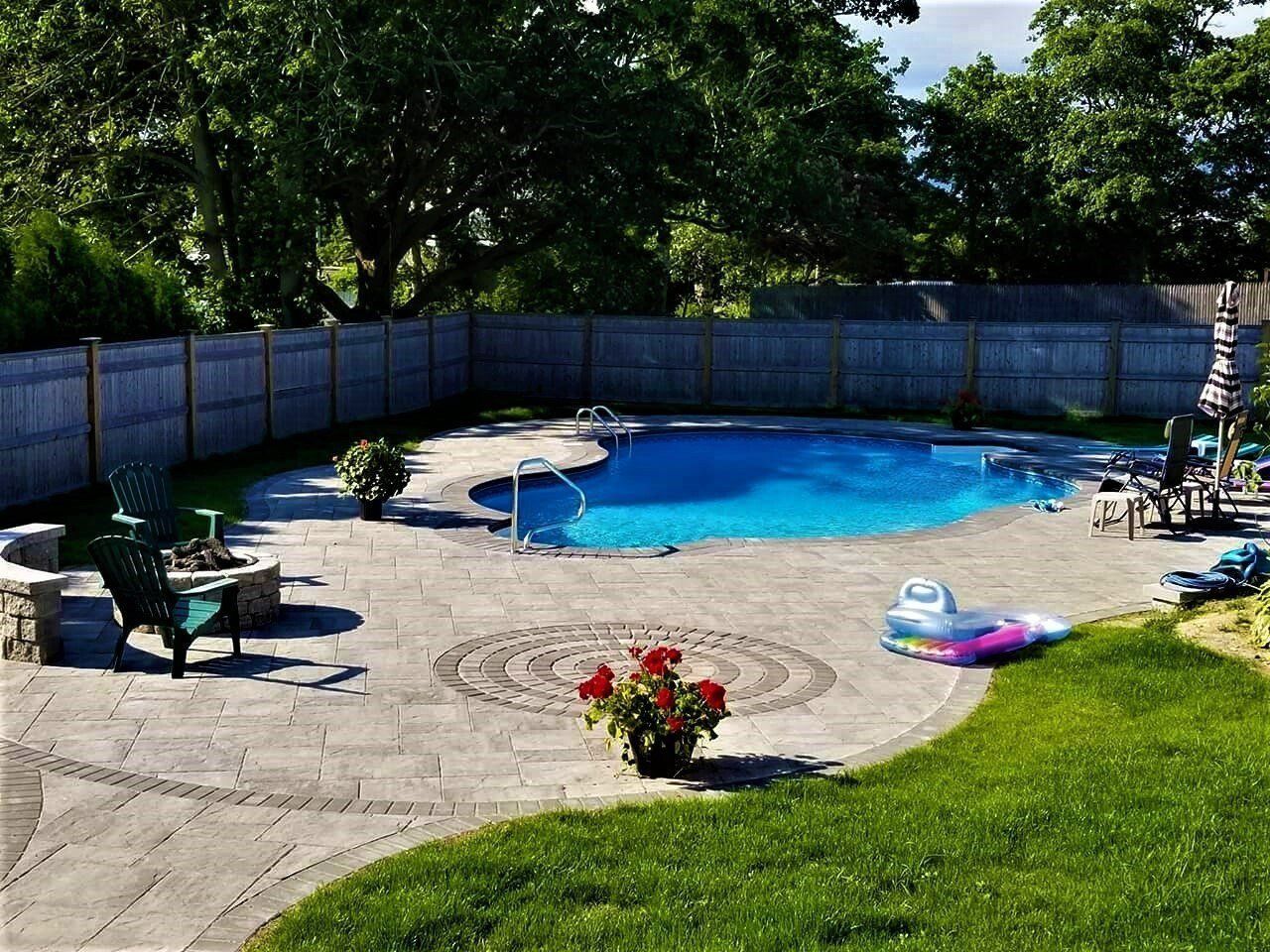 A curved backyard swimming pool with a blue tiled edge, surrounded by a stone patio, potted flowers, and a wooden fence.