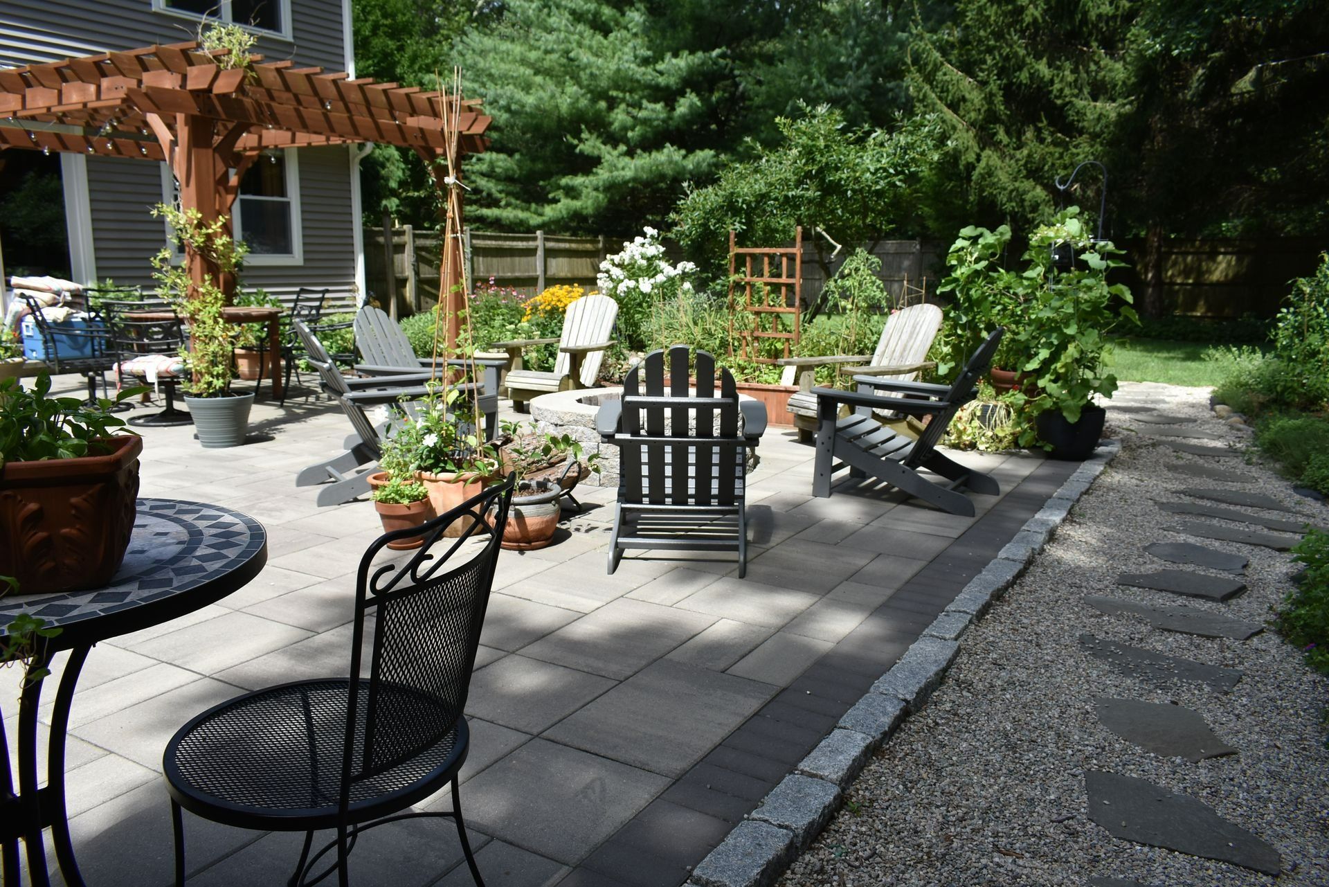A stone patio with a wooden pergola, outdoor chairs, and a gravel path leading through a lush garden.
