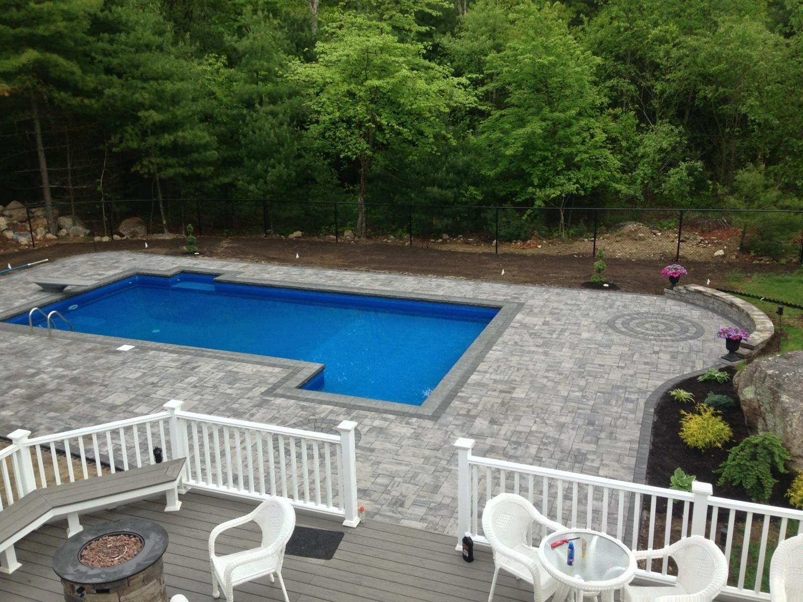 An elevated view of a rectangular swimming pool surrounded by a grey paver patio, with a deck and fire pit in the foreground.