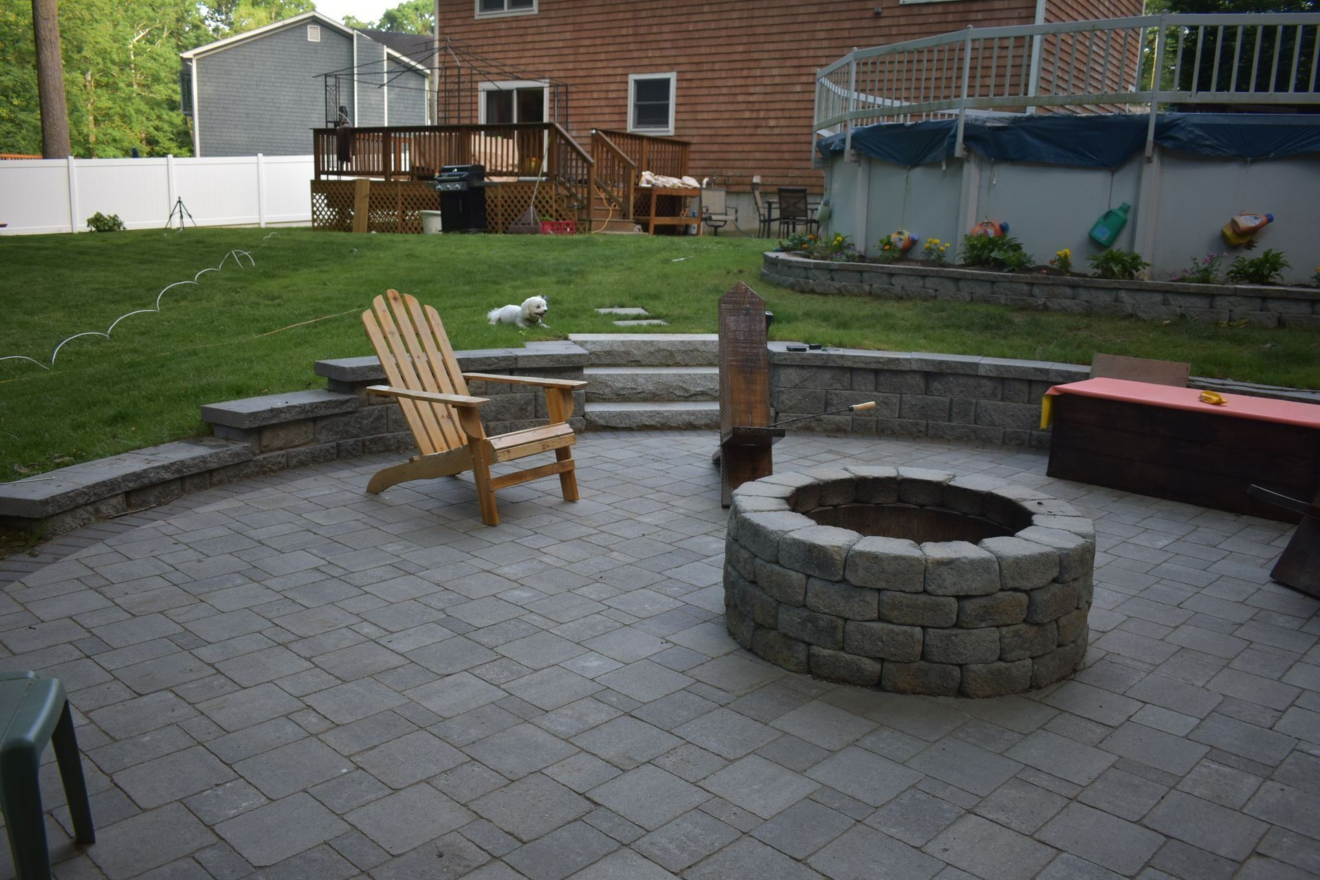 A stone fire pit sits on a gray paver patio in a backyard with a wooden chair and a tiered retaining wall.