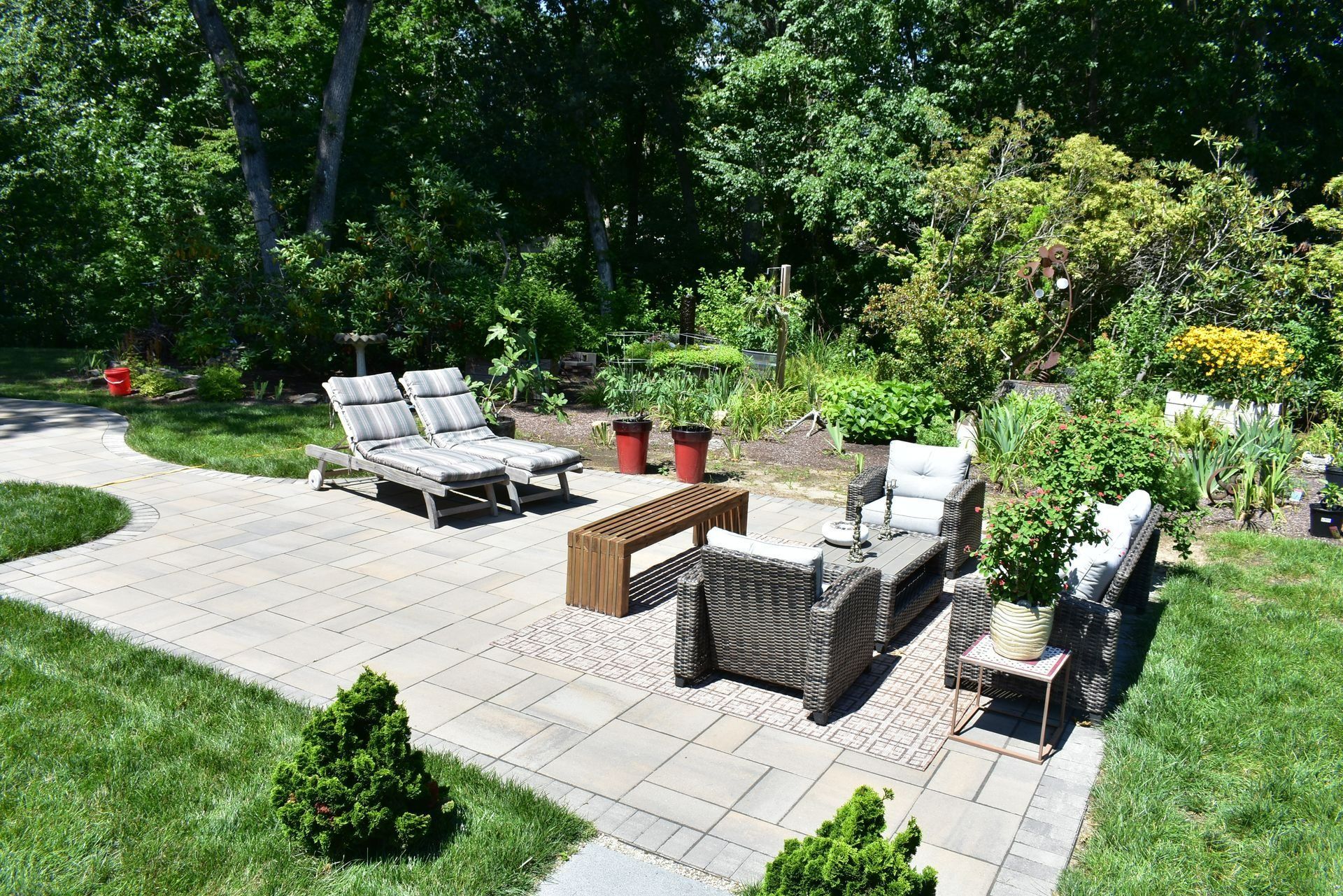 A stone patio in a lush backyard features lounge chairs, wicker armchairs, a wooden bench, and a table in a garden setting.