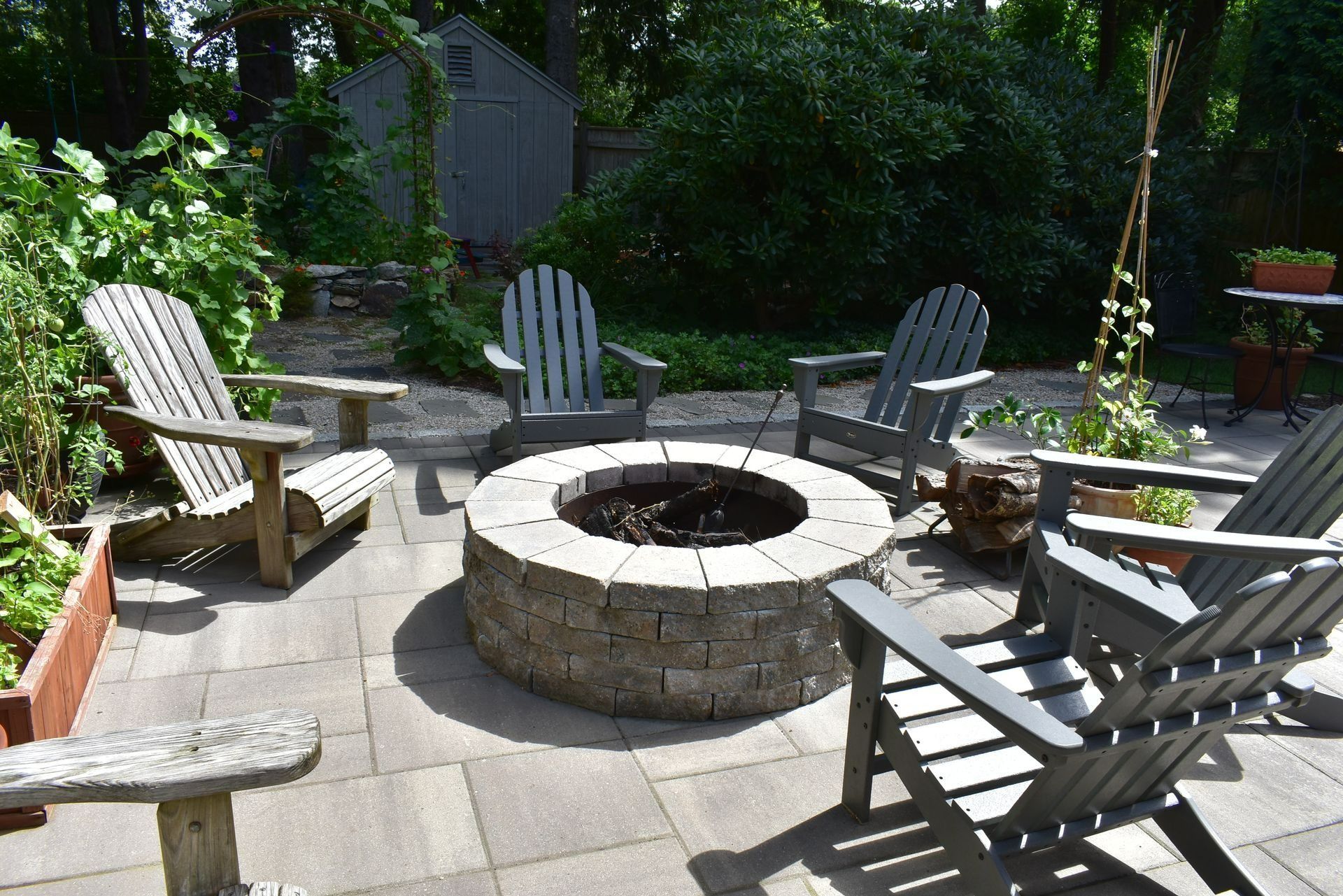 A backyard patio with four gray Adirondack chairs arranged around a circular stone fire pit on a paved surface.