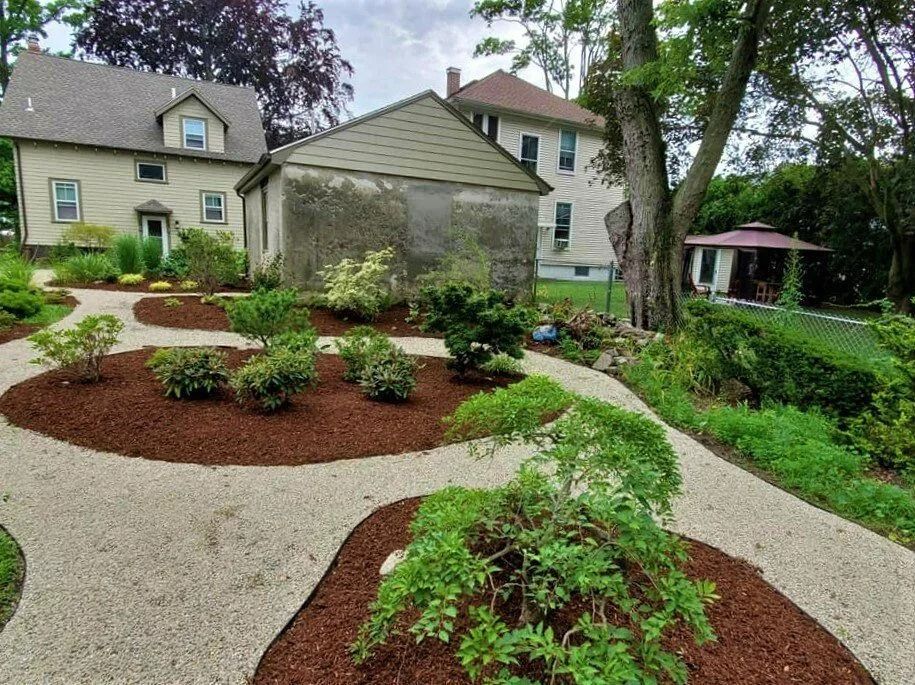 A gravel path winds through a backyard with landscaped mulch beds, various shrubs, two houses, and a small gazebo.