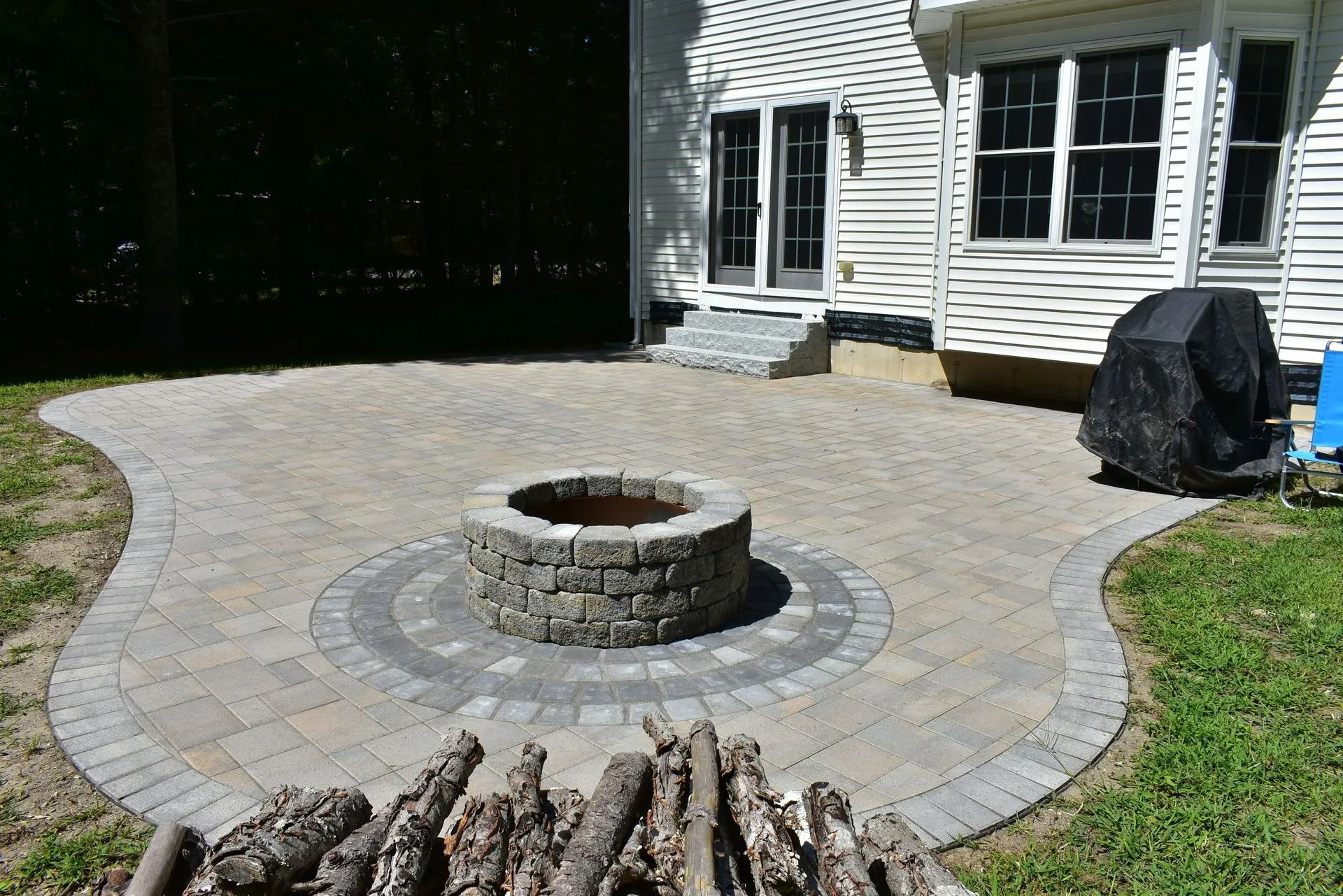 A stone patio with a circular fire pit in the center, next to a house with white siding and a covered grill.