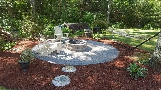 A fire pit with white Adirondack chairs on a stone patio in a yard, surrounded by mulch and lush trees.