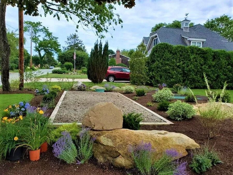 A landscaped yard featuring a central rectangular gravel area, large decorative rocks, and various green and flowering plants.