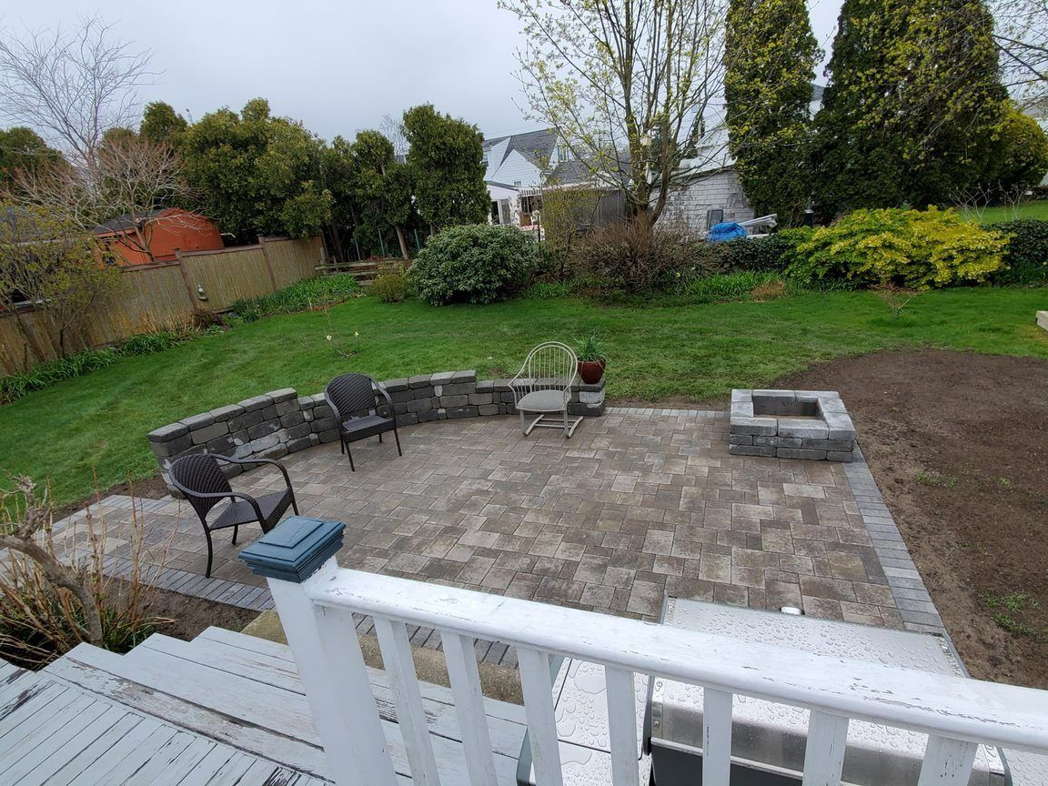 A paved patio with a stone fire pit, seating wall, and two chairs in a backyard viewed from a white wooden deck.