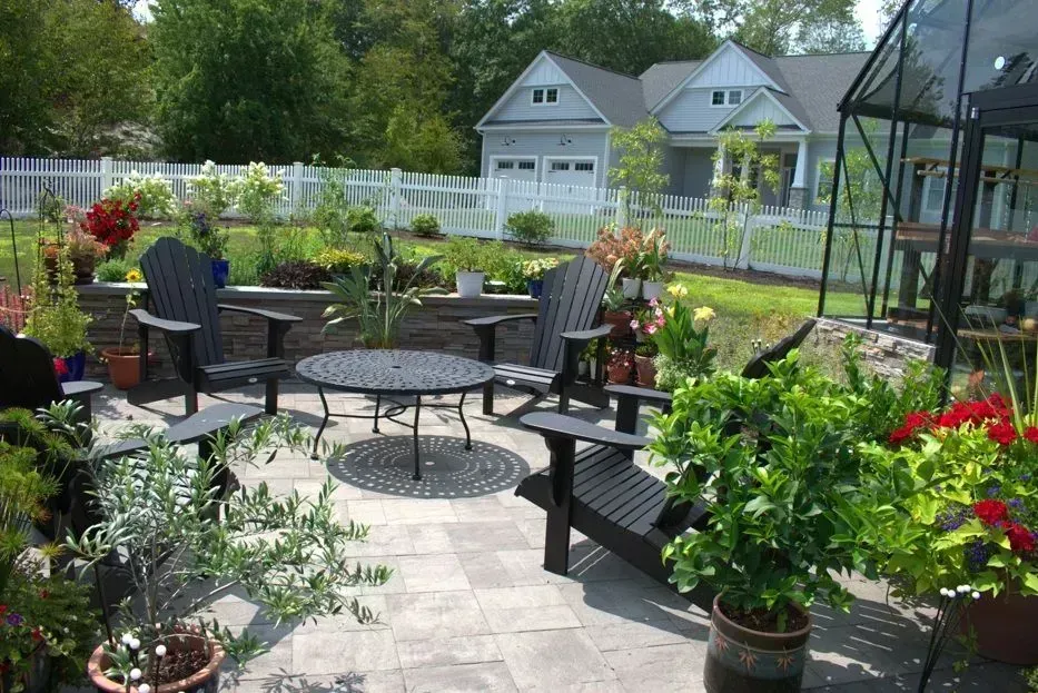 A curved backyard swimming pool with a blue tiled edge, surrounded by a stone patio, potted flowers, and a wooden fence.