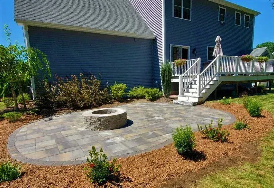 An elevated view of a rectangular swimming pool surrounded by a grey paver patio, with a deck and fire pit in the foreground.