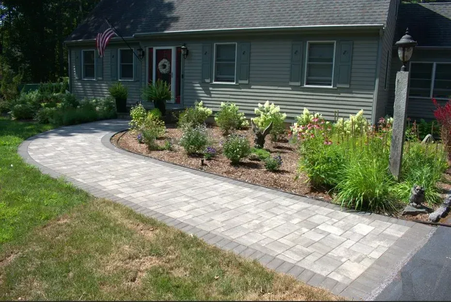 A stone fire pit sits on a gray paver patio in a backyard with a wooden chair and a tiered retaining wall.