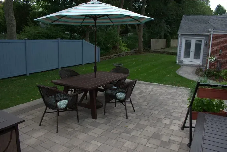 A stone patio in a lush backyard features lounge chairs, wicker armchairs, a wooden bench, and a table in a garden setting.