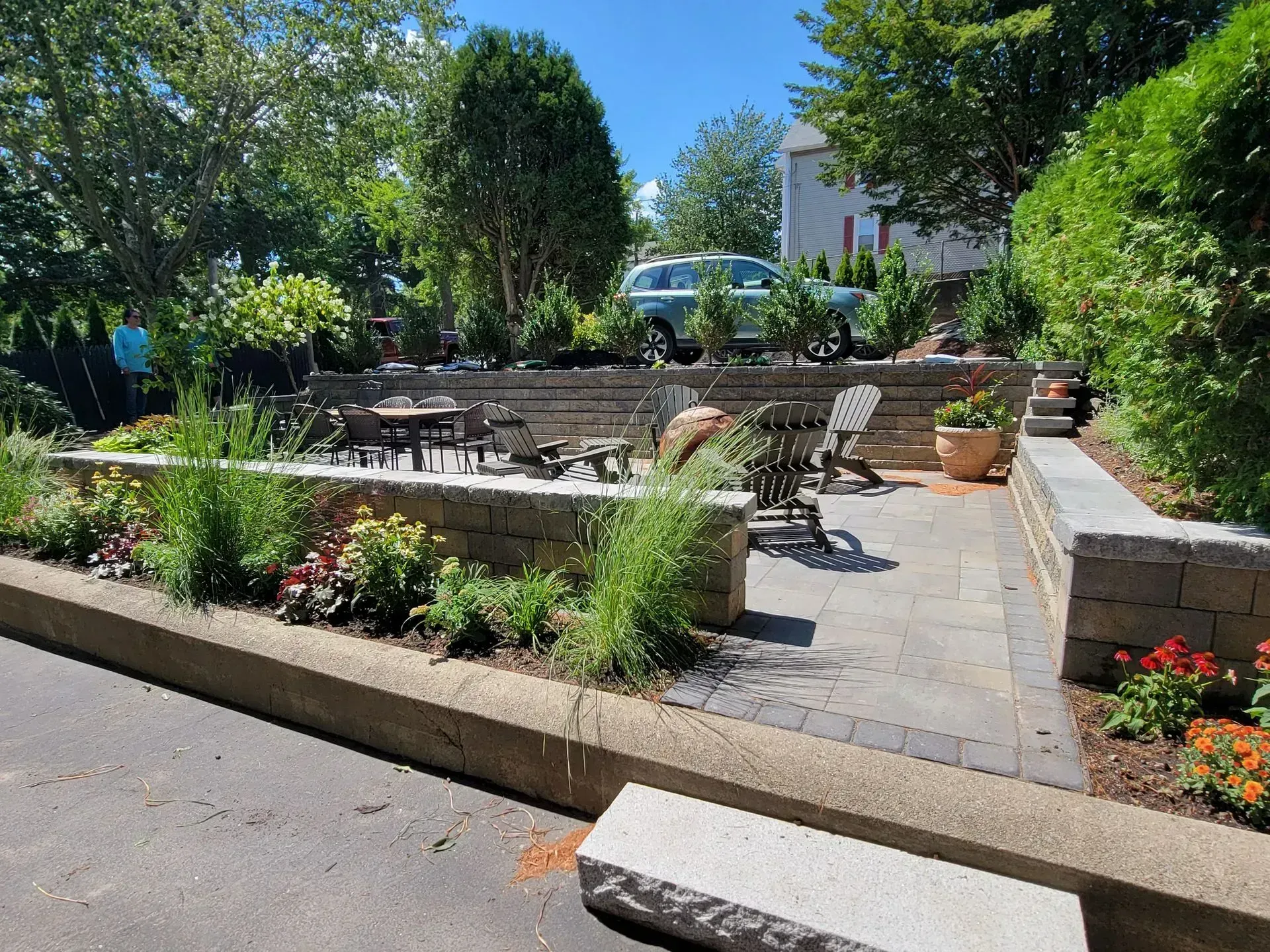 A fire pit with white Adirondack chairs on a stone patio in a yard, surrounded by mulch and lush trees.