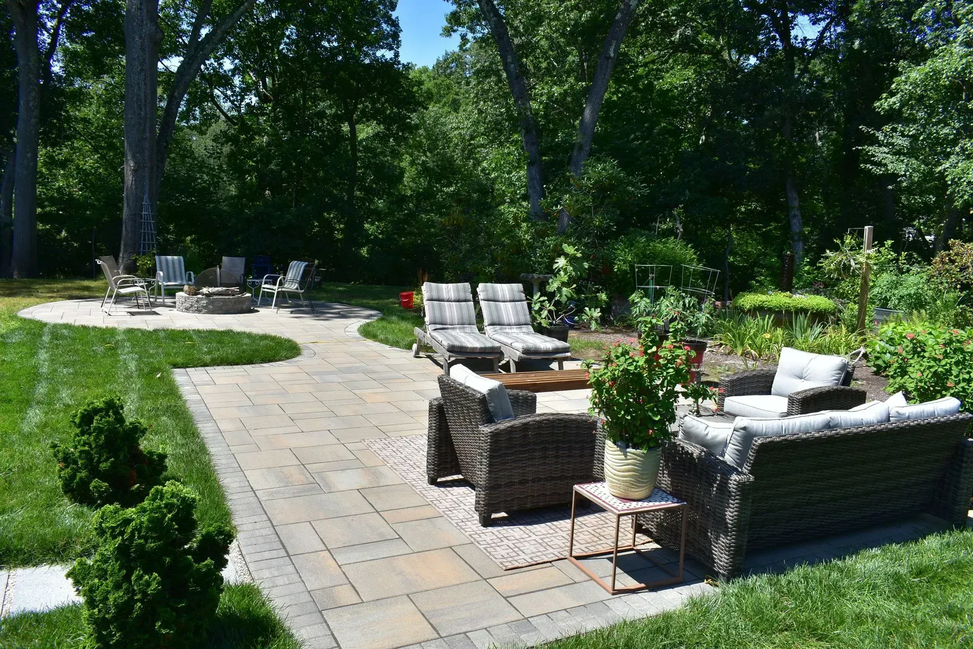 A paved patio with a stone fire pit, seating wall, and two chairs in a backyard viewed from a white wooden deck.