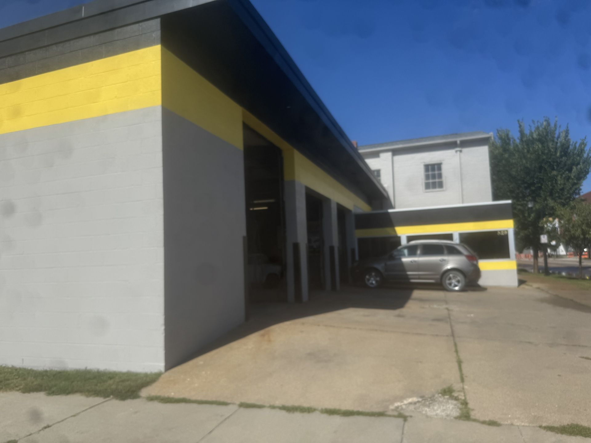 Gray building with yellow stripe, covered walkway, and a parked car in a sunny lot