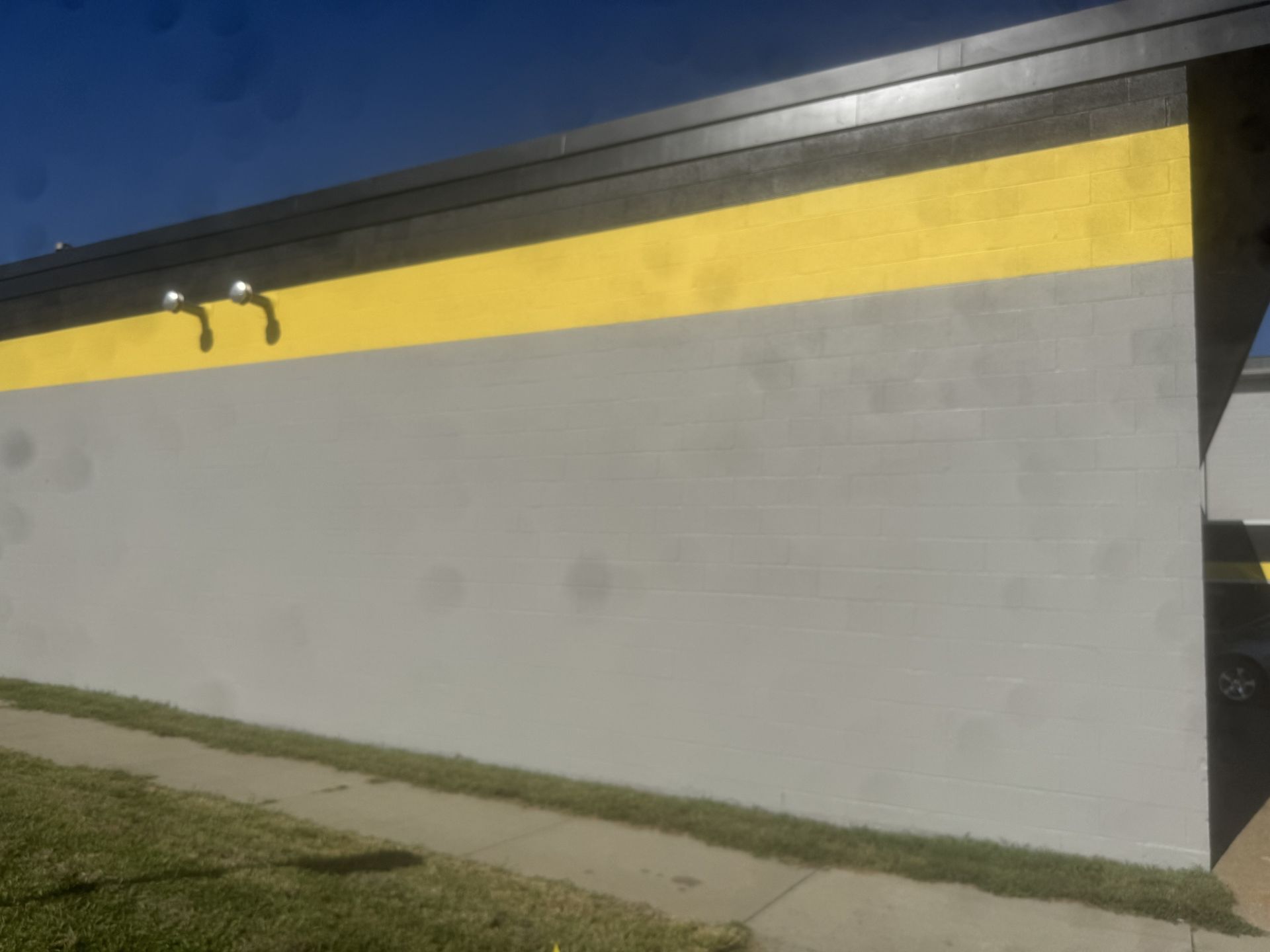Gray building wall with a yellow stripe near the roof and small lights, beside a grassy strip under a blue sky