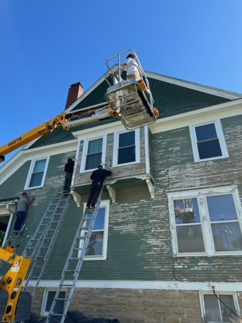 People painting a two-story house with peeling green paint, using a lift and ladders, under a blue sky.