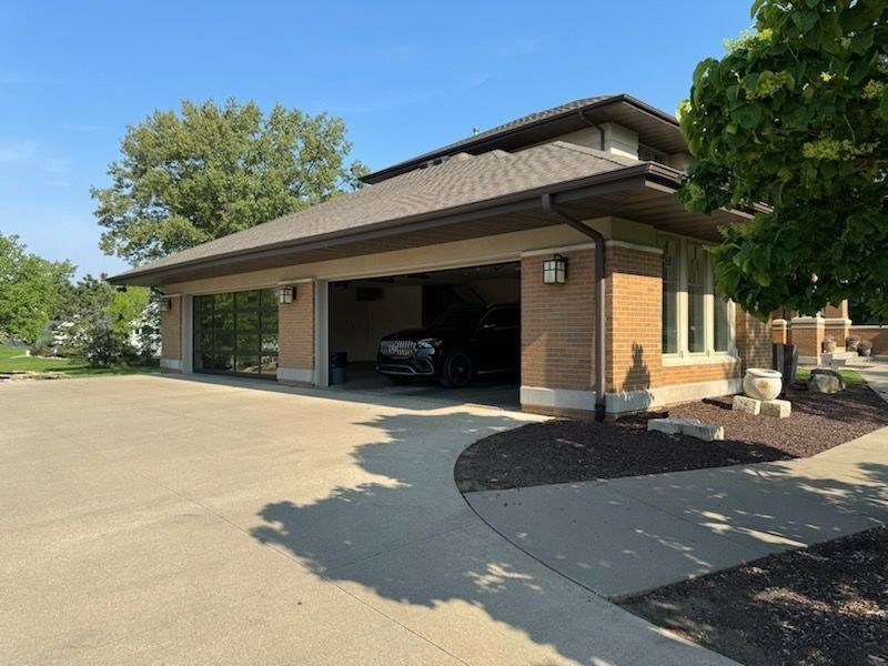 Garage with open doors; a car inside, brick exterior, concrete driveway, and a green tree in the background.