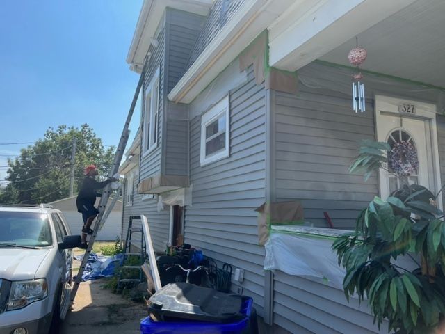 Person on ladder working on the side of a two-story house, partially covered with gray siding.